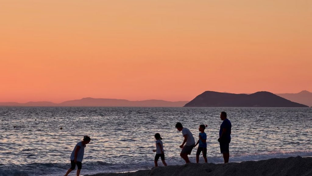 A family enjoying a sunset by the beach at Perdika, Greece, with orange skies and serene waves.