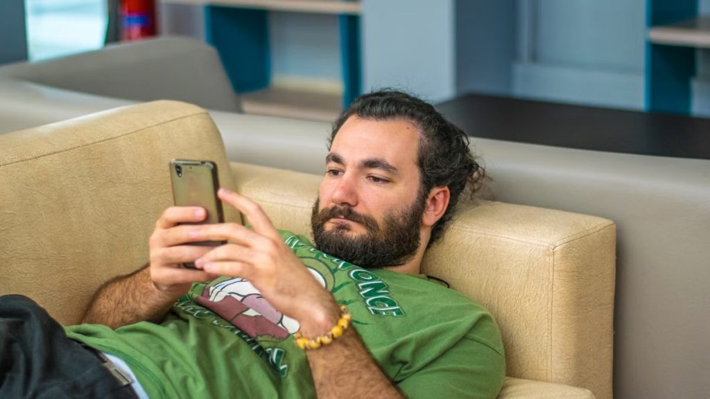 Bearded man leisurely laying on a sofa, engrossed in smartphone indoors.