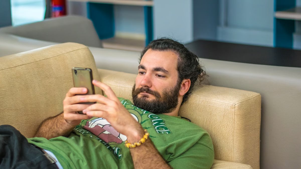 Bearded man leisurely laying on a sofa, engrossed in smartphone indoors.